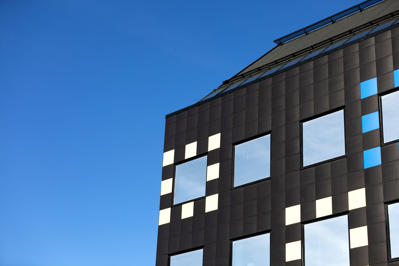 Fasad på en modern byggnad med mörka paneler och rutmönster i vitt och blått mot en klarblå himmel